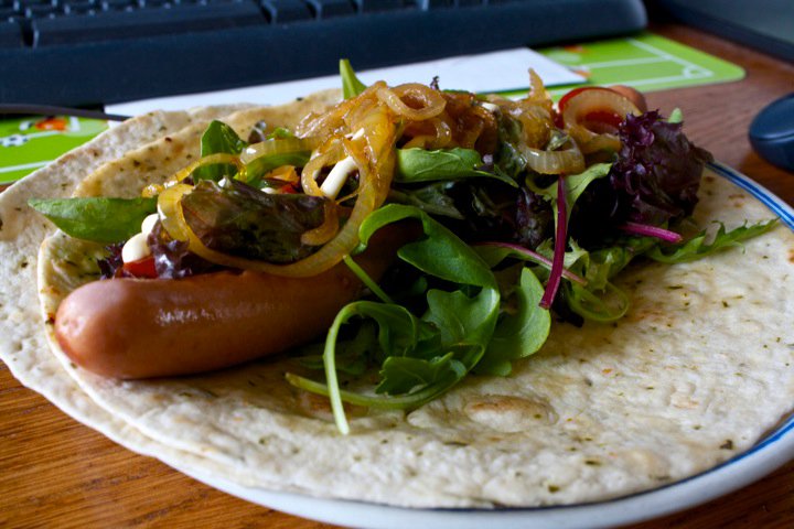 A hotdog tortilla wrap with plenty of greens and condiments.